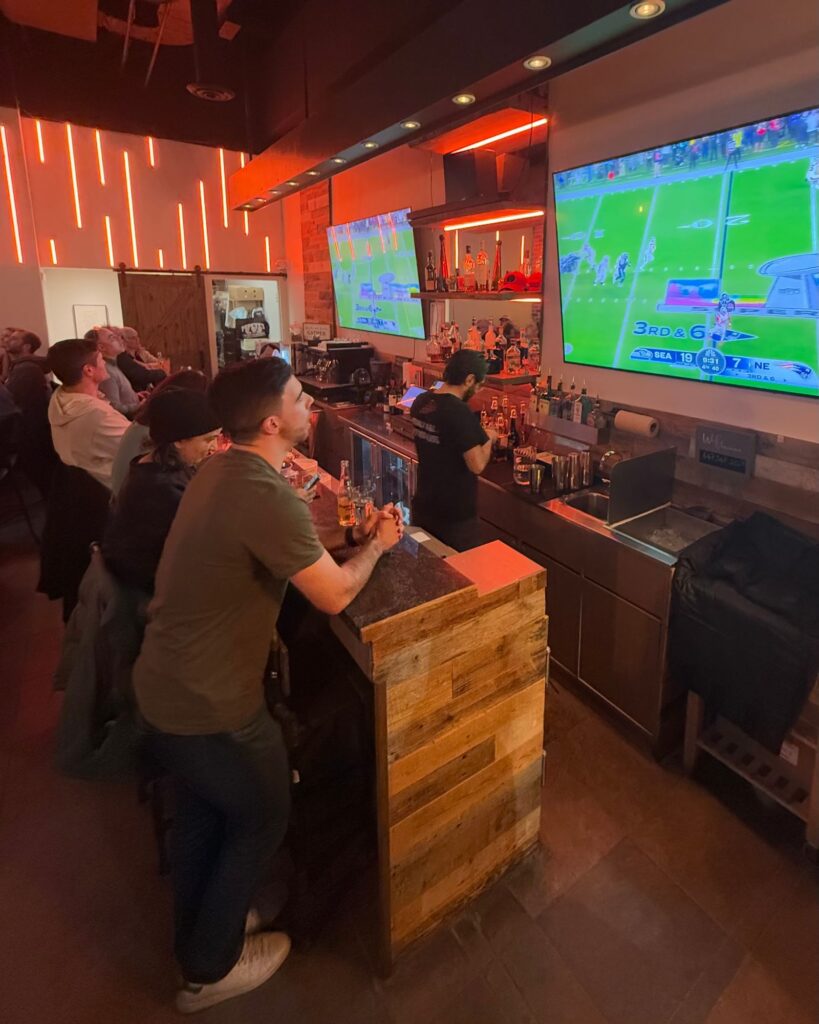 Guests at the JZ's Pizza bar watching a football game on two large wall-mounted TVs, with orange neon light strips on the wall