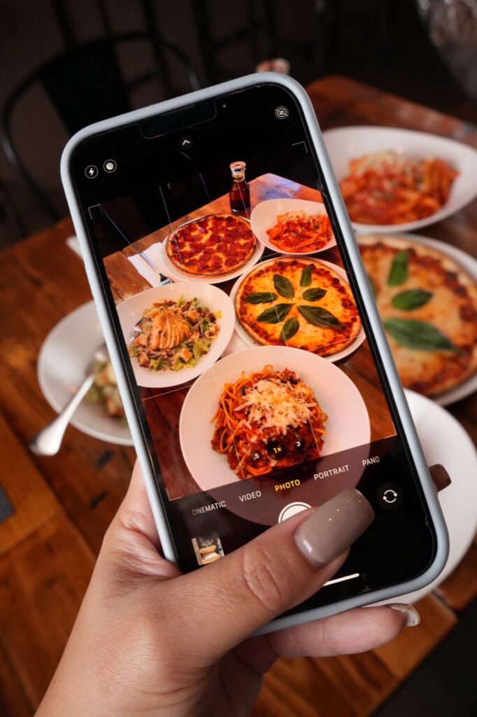 Phone camera framing a table of JZ's Pizza dishes: pepperoni and margherita pizzas, penne, a chicken Caesar salad and spaghetti Bolognese